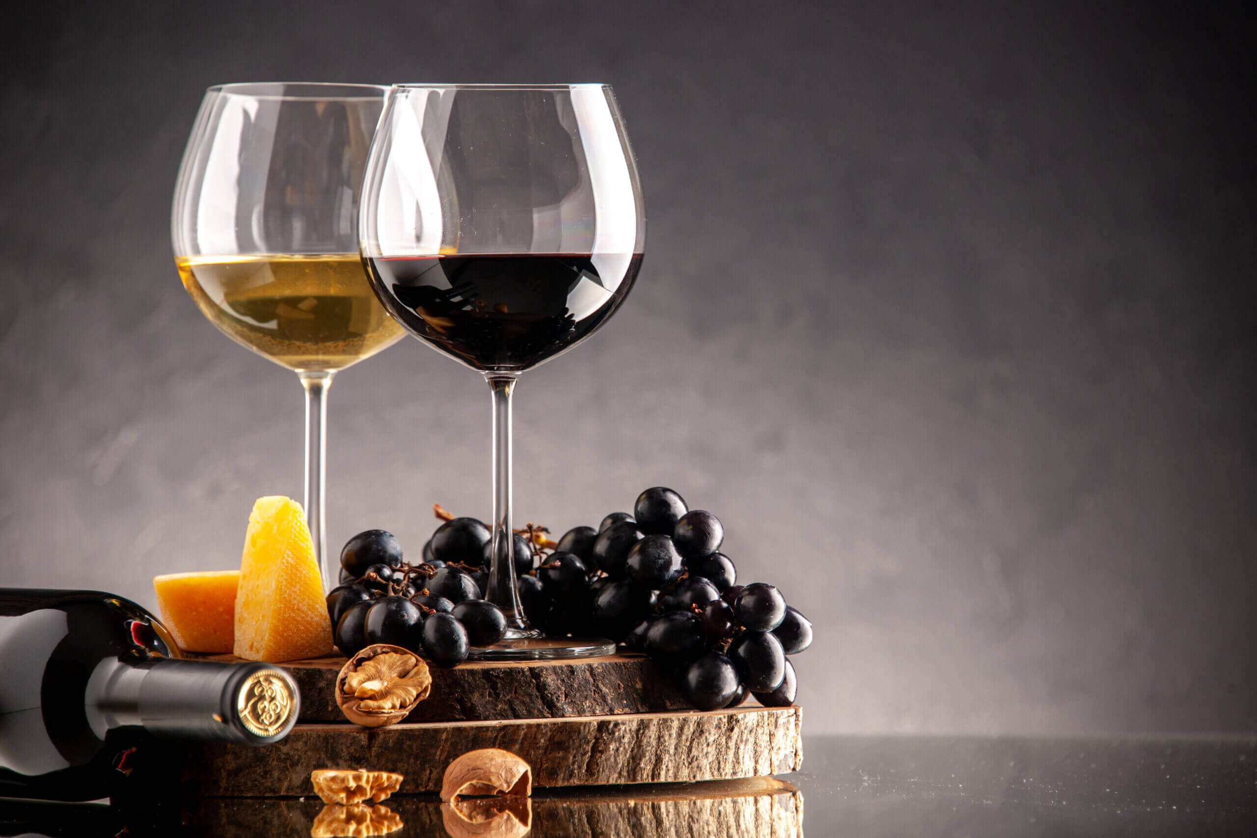 front view wine glasses fresh grapes walnuts yellow cheese wood board overturned bottle dark background scaled